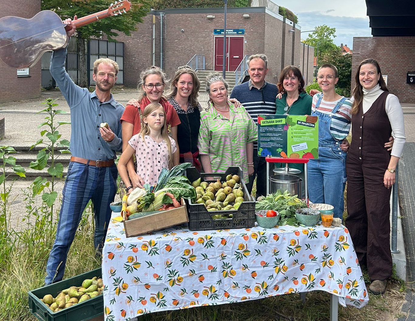 Vrijwilligers bij een kraam met geoogste groenten; iemand houdt een gitaar omhoog